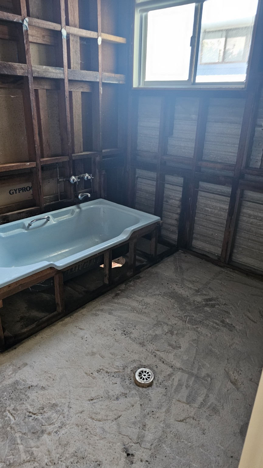 Bathroom with wall linings removed to expose framing, with a bathtub retained and the floor stripped.