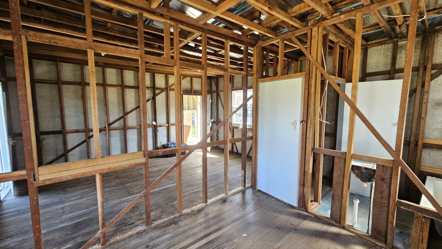 Room with wall and ceiling linings removed, leaving timber framing and floorboards visible.