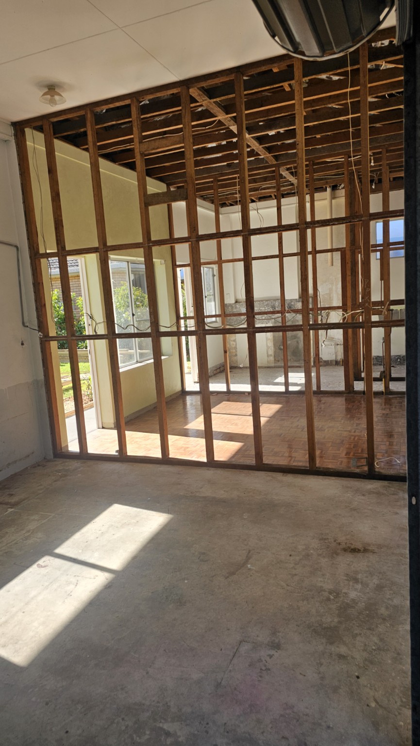 Room with wall and ceiling linings removed to expose timber framing, viewed through an internal wall frame.