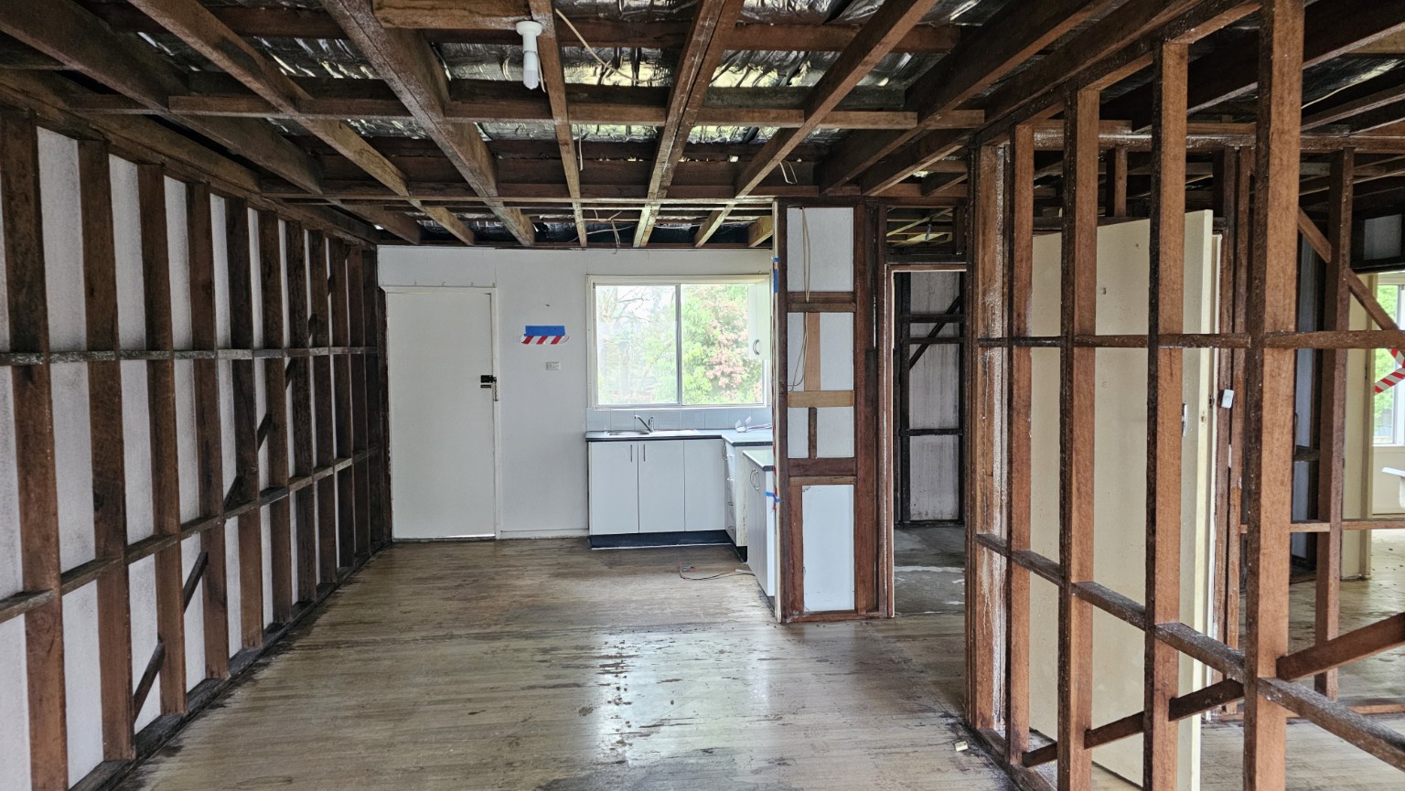 Internal wall and ceiling linings removed to expose framing beside a retained kitchenette and timber floor.